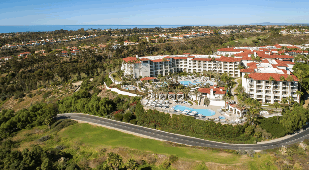 Aerial view of a luxury coastal resort with pools, palm trees, and red-tile roofs surrounded by greenery.