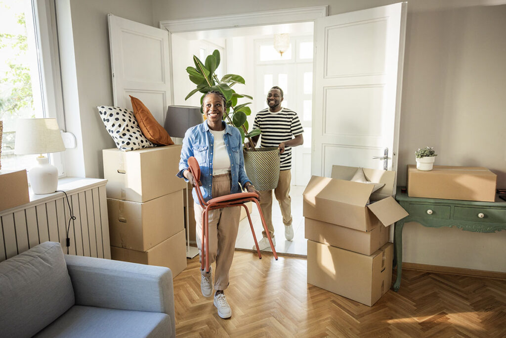 Couple carrying boxes and a chair while moving into a new home.