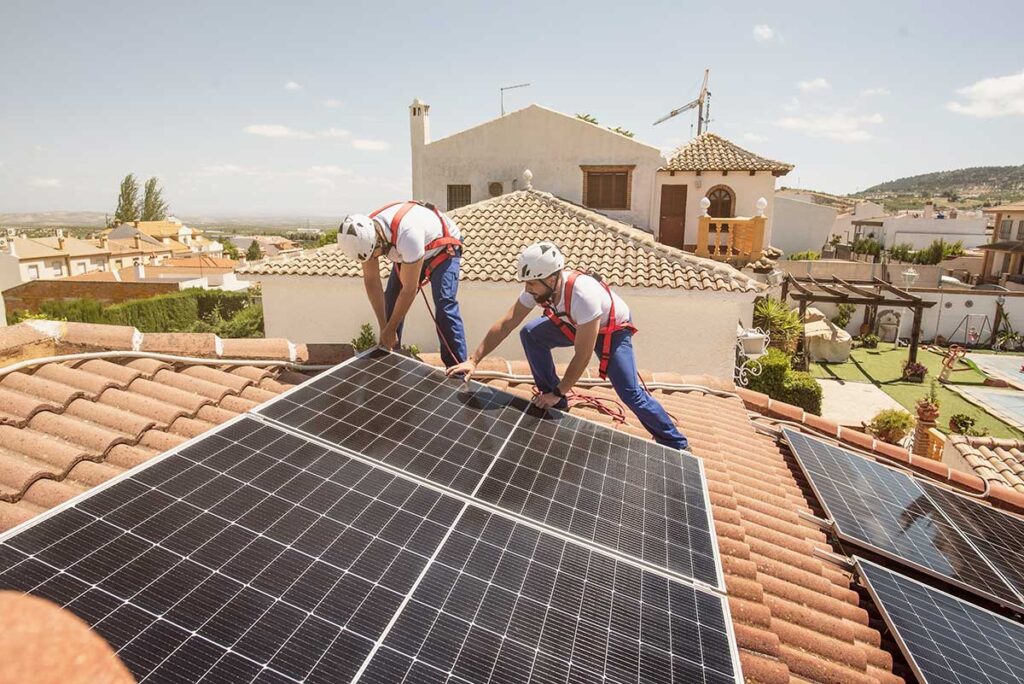 Two installers place solar panels on a tiled residential roof in a sunny neighborhood.