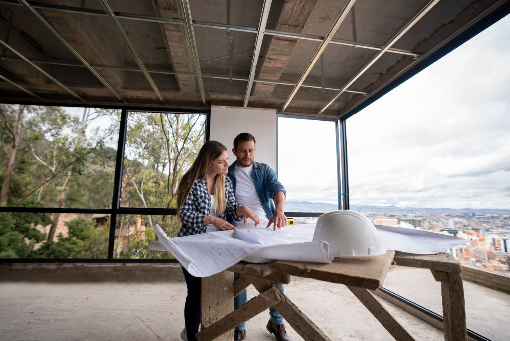 Couple reviewing home construction plans inside a partially completed modern house with large windows and open interior framing.